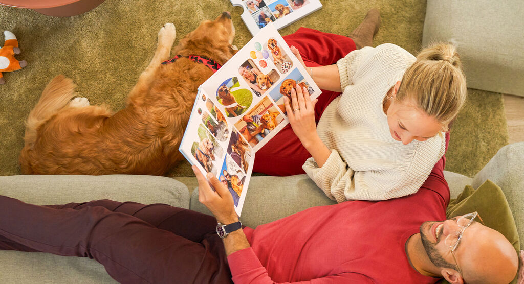 Eine Frau sitzt mit einem Hund am Boden, ein Mann liegt auf dem Sofa. Gemeinsam blättern sie durch ein CEWE FOTOBUCH.