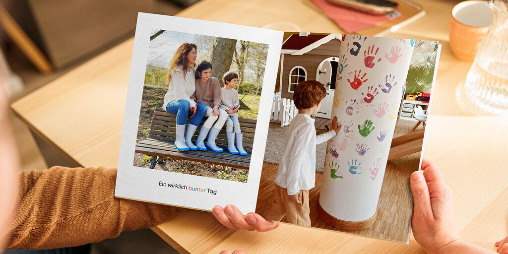 Zwei Kinder und ein Jugendlicher sitzen auf einer Holzbank und schauen neugierig in ein Fotobuch, das eine Darstellung bunter Handabdrücke zeigt