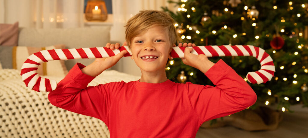 Ein fröhliches Kind hält einen großen Zuckerstangen-Lutscher und zeigt einen glücklichen Gesichtsausdruck. Im Hintergrund strahlt ein Weihnachtsbaum mit Lichtern.