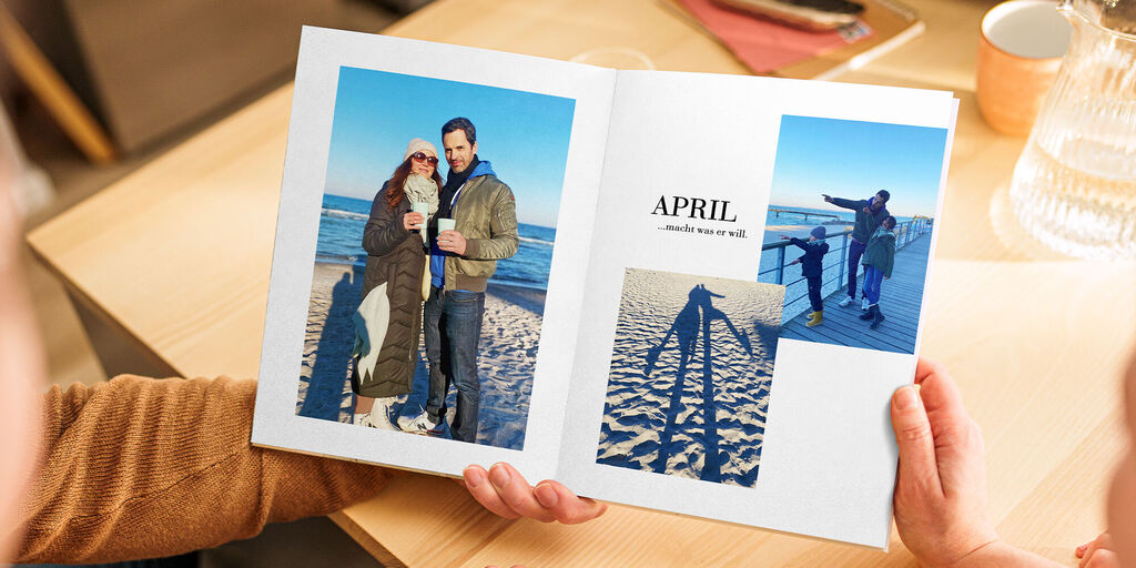 Zwei Personen blättern in einem CEWE Fotobuch mit Erinnerungen an den Urlaub im April, darunter Fotos am Strand und bei Sonnenuntergang.