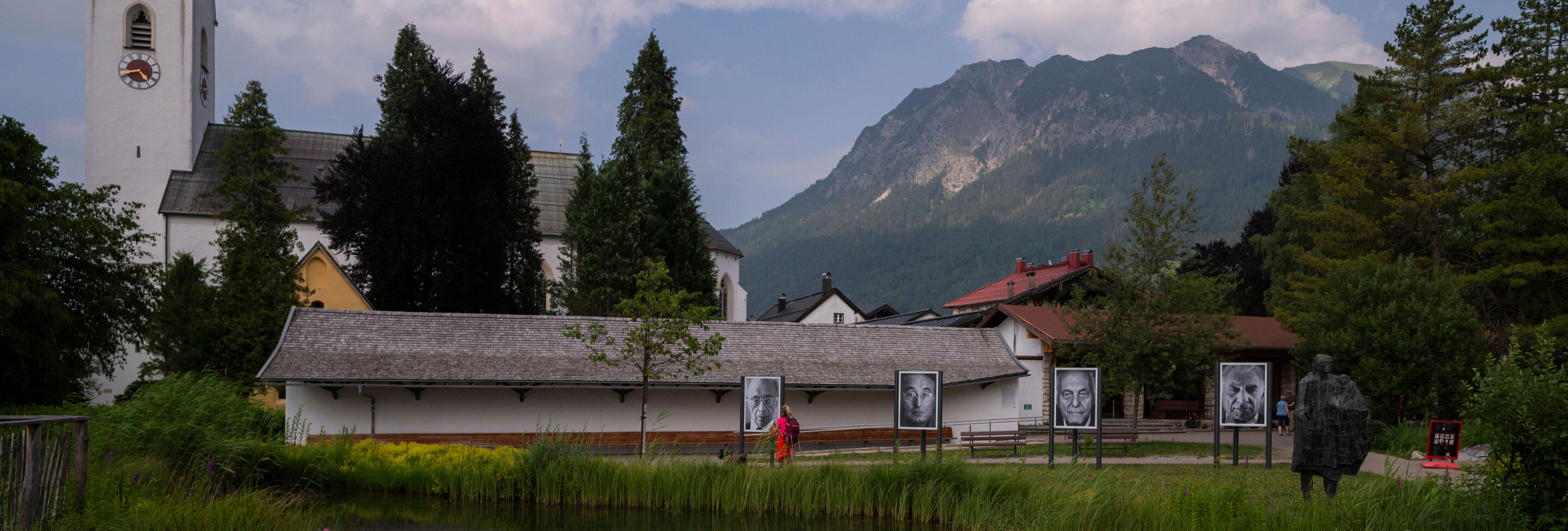 Auf einer grünen Wiese stehen vier große Schwarz-weiß-Fotografien von verschiedenen Menschen. Im Hintergrund sind eine Kirche und ein Berg.