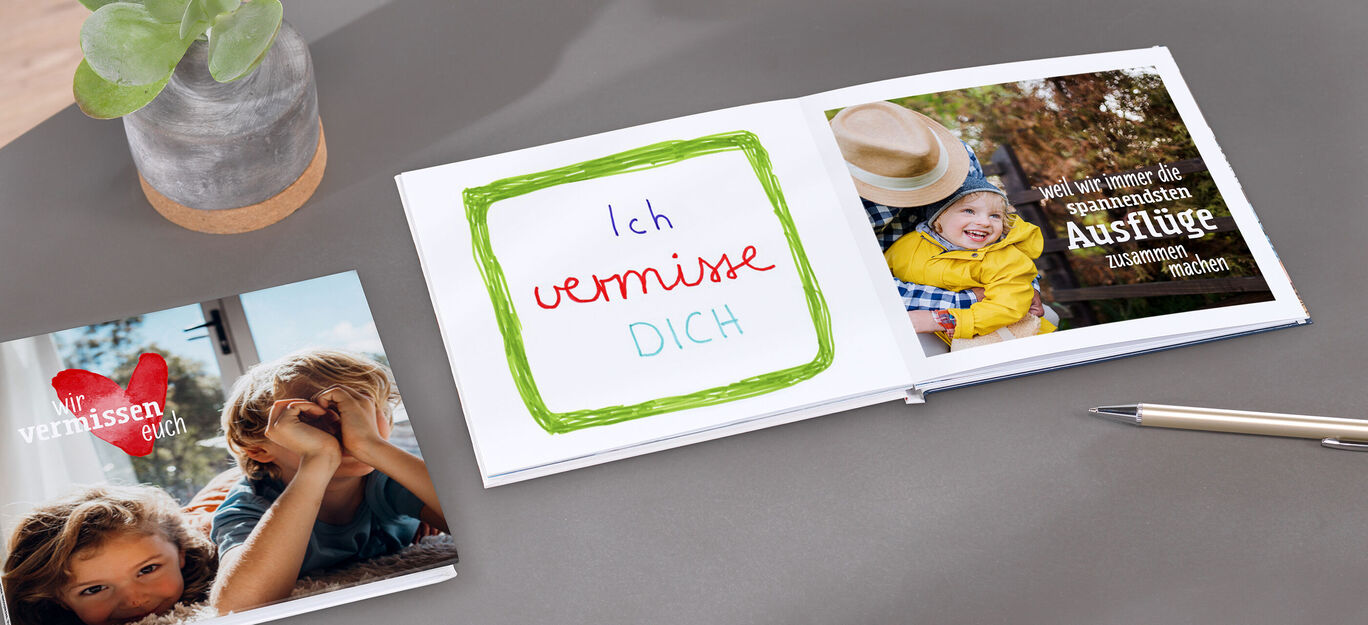 Auf einem Tisch stehen ein Fotobuch mit Herz-Motiv und ein offenes CEWE FOTOBUCH, das eine liebevolle Botschaft zeigt. Eine Pflanze und ein Stift sind daneben.