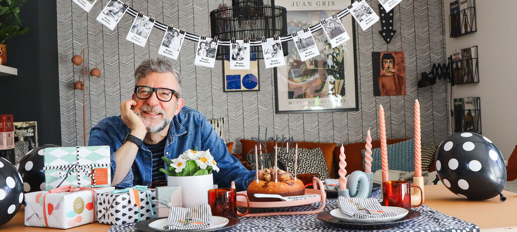 Ein Mann sitzt an einem festlich gedeckten Tisch mit Geschenken, Luftballons, Kerzen und Geburtstagsdekoration in einem Wohnzimmer.