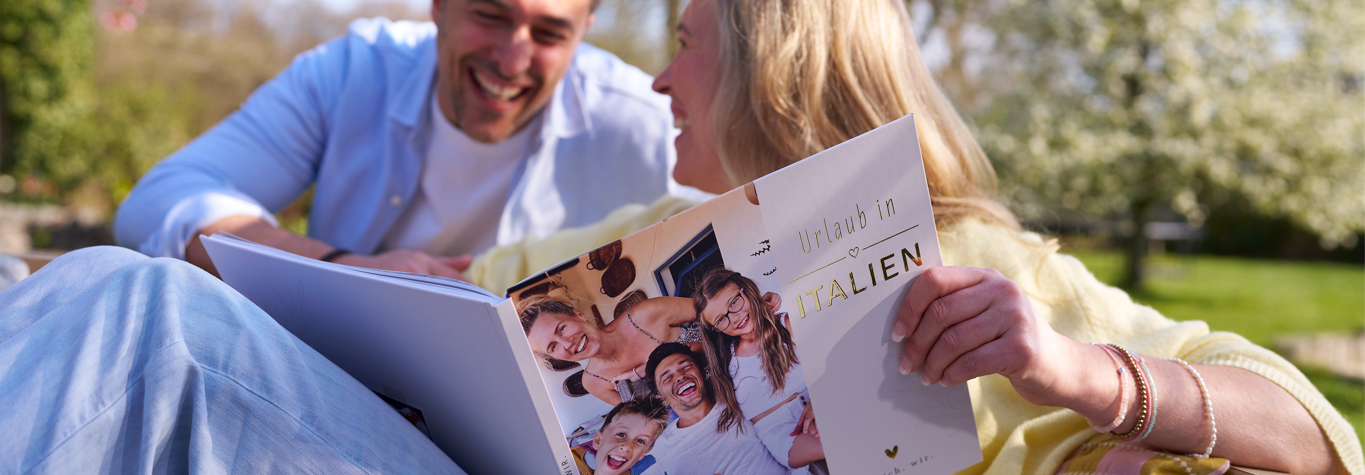 Zwei Personen sitzen draußen und blättern gemeinsam in einem Fotobuch mit Familienbildern.