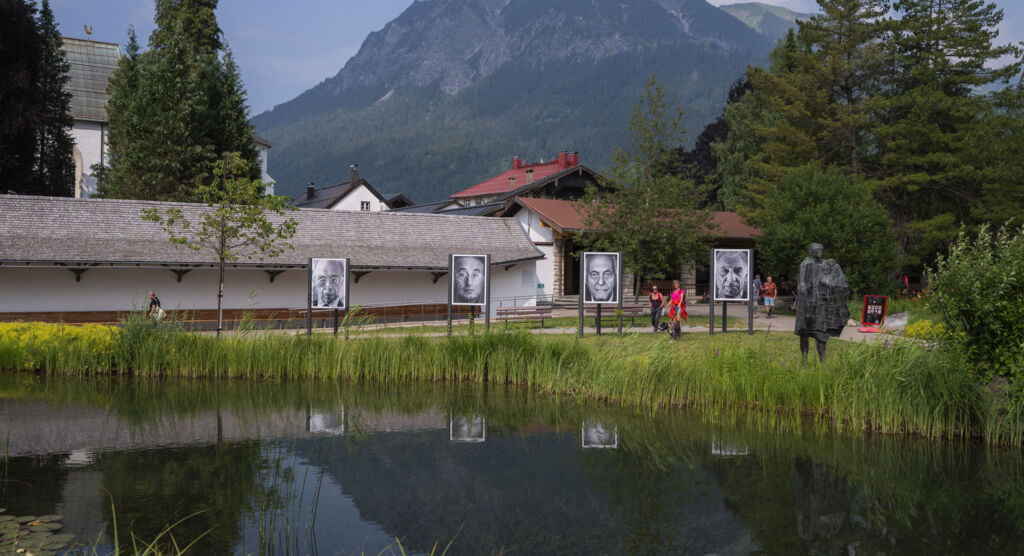An einem See findet eine Fotoausstellung mit großen Porträts statt. Im Hintergrund ist ein Berg zu sehen.