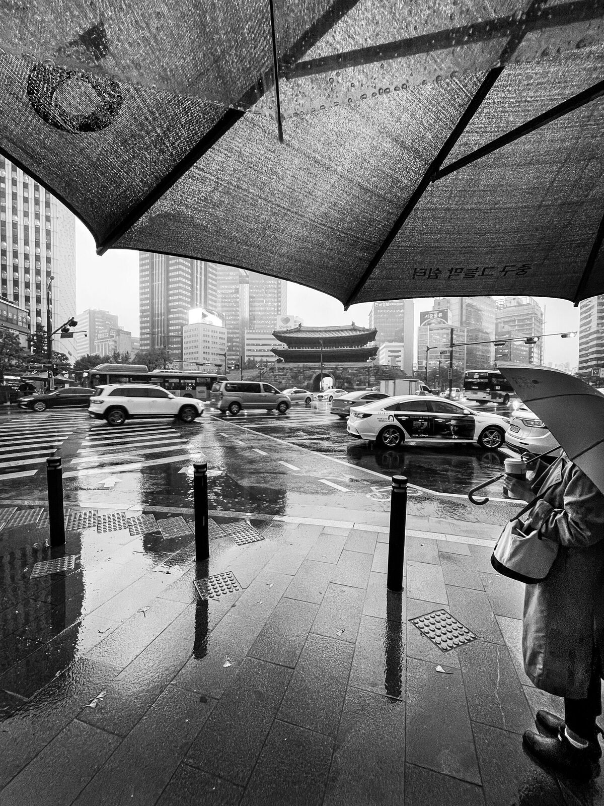Eine Person in einem Mantel, die unter einem großen Regenschirm steht, während es regnet. Im Hintergrund sind hochmoderne Gebäude und Autos zu sehen.