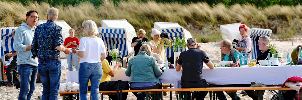 Mehrere Menschen sitzen an einem langen Tisch am Strand und unterhalten sich.