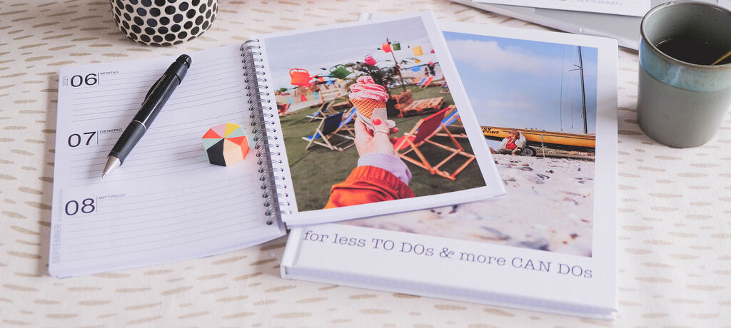 Ein Notizbuch mit Fotos von einem Sommerurlaub. Eine Person isst ein Eis und hält die Kamera. Bunte Gegenstände liegen auf dem Tisch.