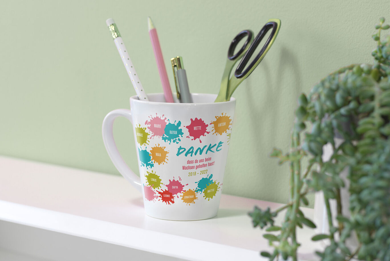 Weißer Becher mit bunten Farbspritzern, gefüllt mit Schreibutensilien wie Stift und Schere