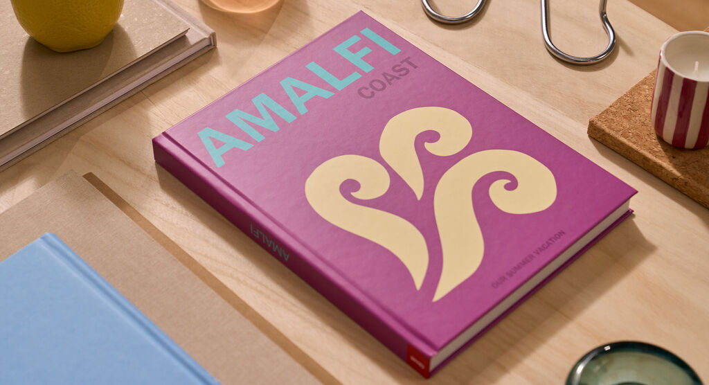 Auf einem Tisch liegt ein CEWE FOTOBUCH. Das Cover ist violett und mit dem Schriftzug „Amalfi“ gestaltet.