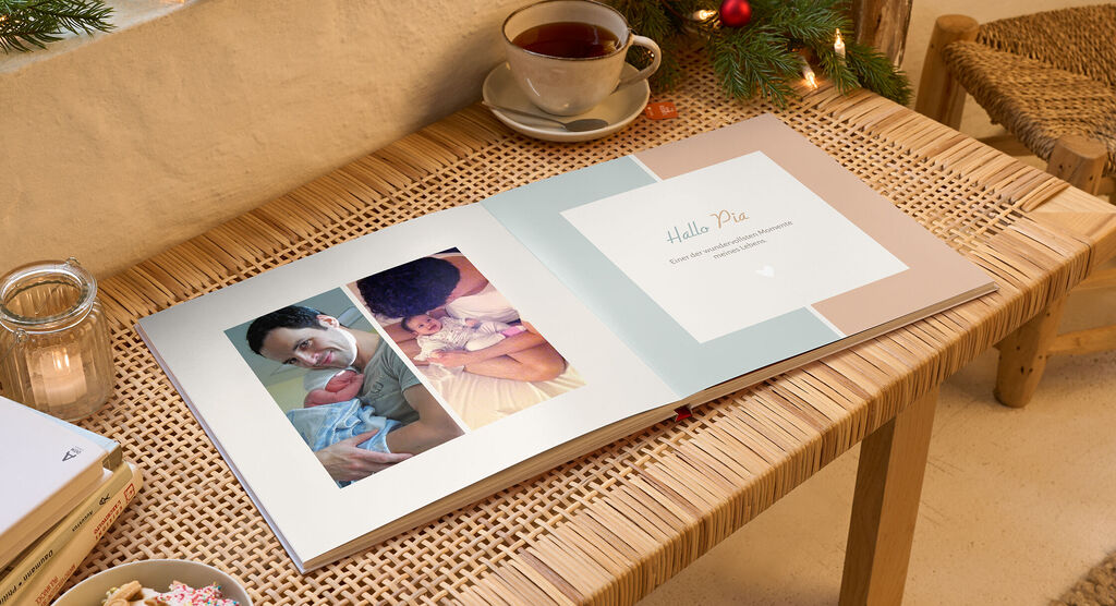 Ein offenes CEWE Fotobuch zeigt einen liebevollen Schnappschuss einer Familie. Auf der linken Seite ist ein Vater mit seinem Baby zu sehen, auf der rechten Seite steht "Hallo Pia" auf einer hellen Hintergrundseite.