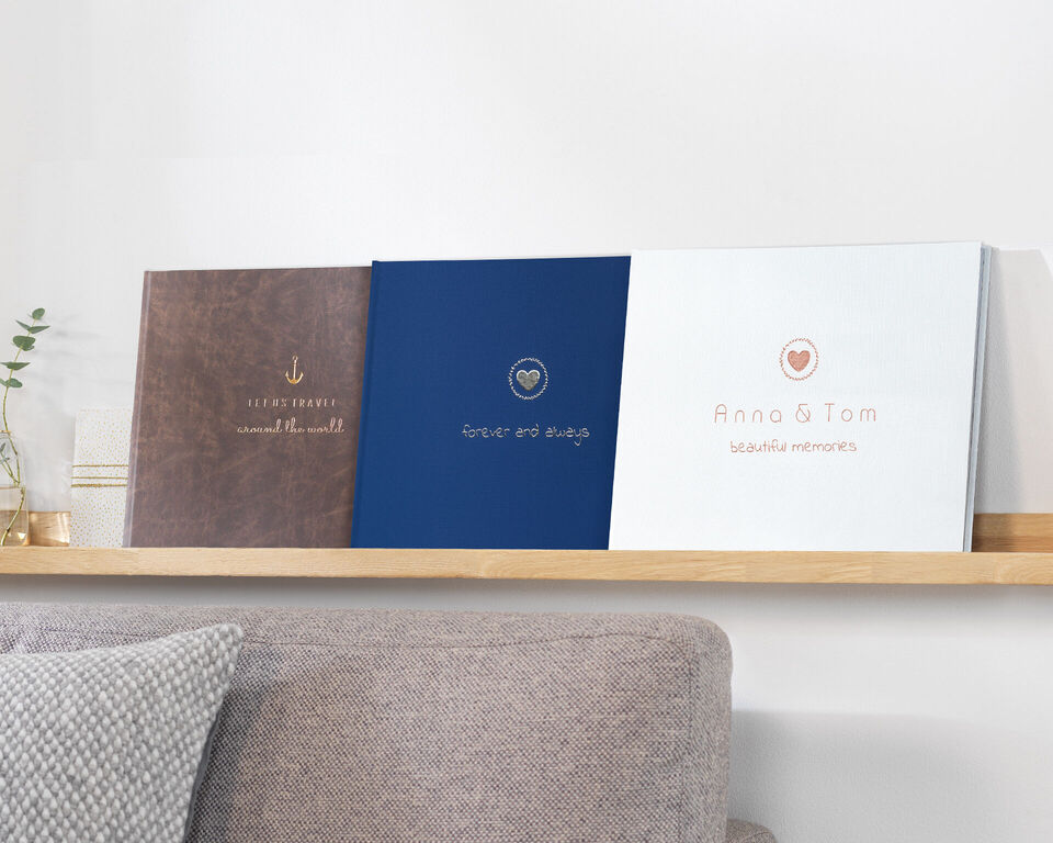 Drei verschiedene Fotobücher stehen auf einem Regal. Sie haben unterschiedliche Cover-Designs in Braun, Blau und Weiß und zeigen liebevolle Titel.