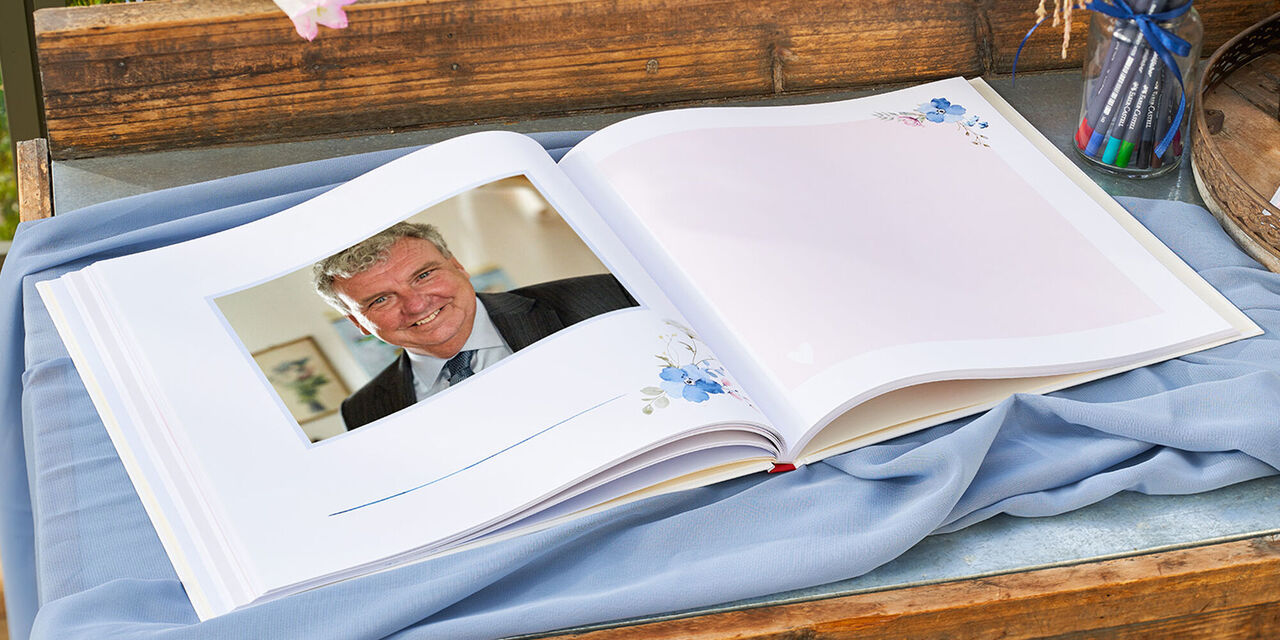 Ein Gästebuch liegt aufgeklappt auf einem Holztisch. Die Doppelseite zeigt links das Foto eines Mannes und rechts einen handschriftlichen Eintrag. Neben dem Buch steht ein Glas mit Stiften. Daneben liegt ein Bogen mit Stickern. Im Hintergrund stehen Blumen.