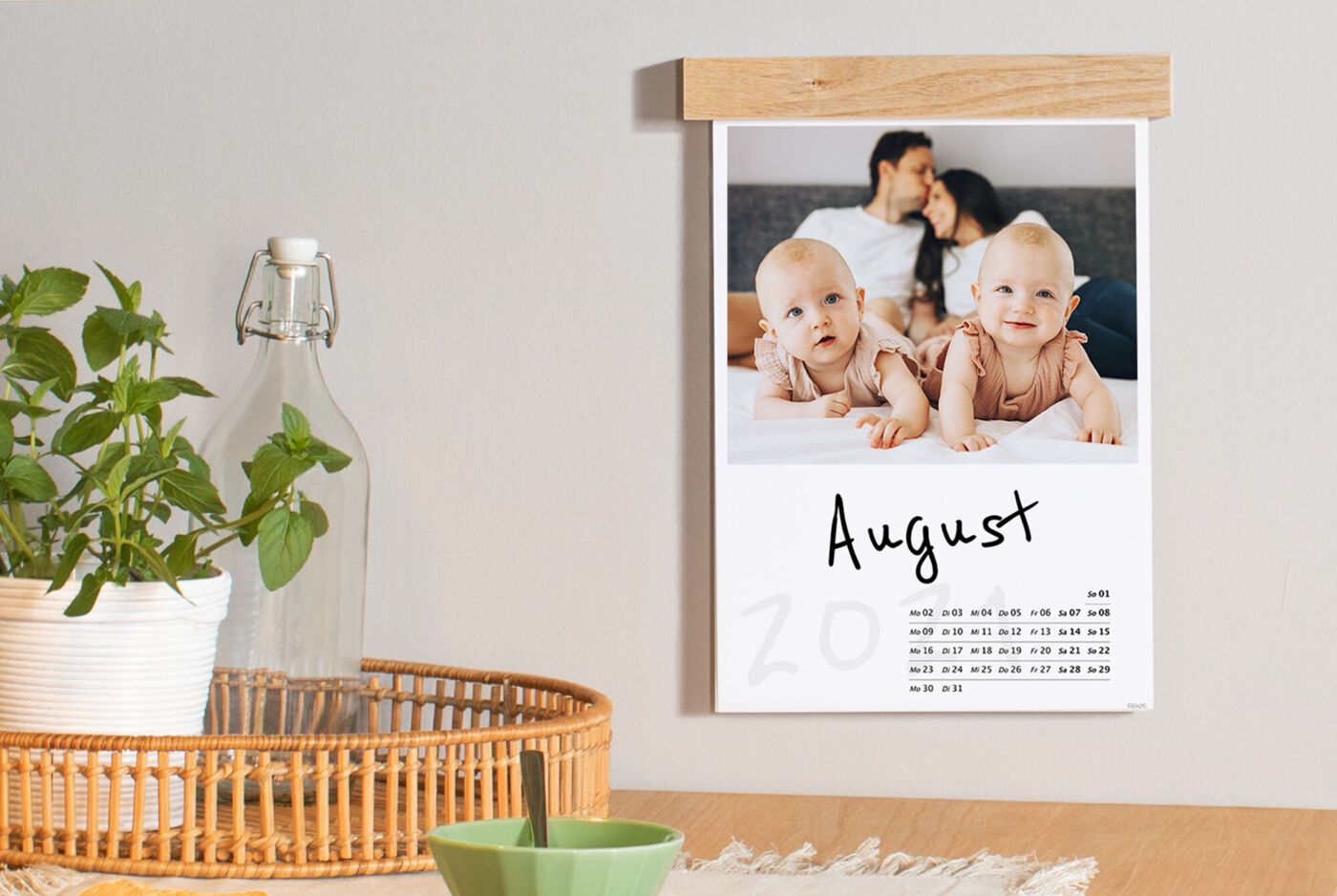 Kalender für August mit zwei Babys, die auf dem Boden spielen. Pflanzen und ein Glas Wasser sind vorhanden.