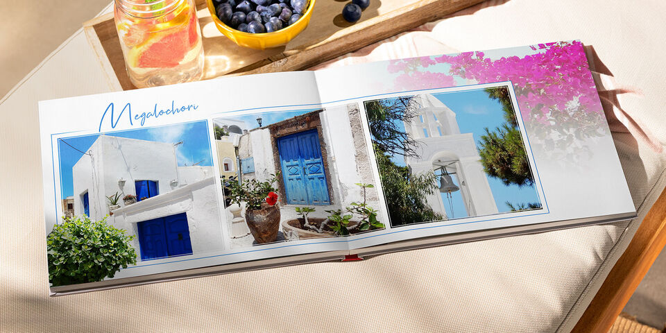 Ein offenes CEWE FOTOBUCH mit Bildern von weißen Häusern und blauen Türen auf einer sonnigen Terrasse in Griechenland. Im Hintergrund blühen bunte Blumen und frisches Obst liegt bereit.