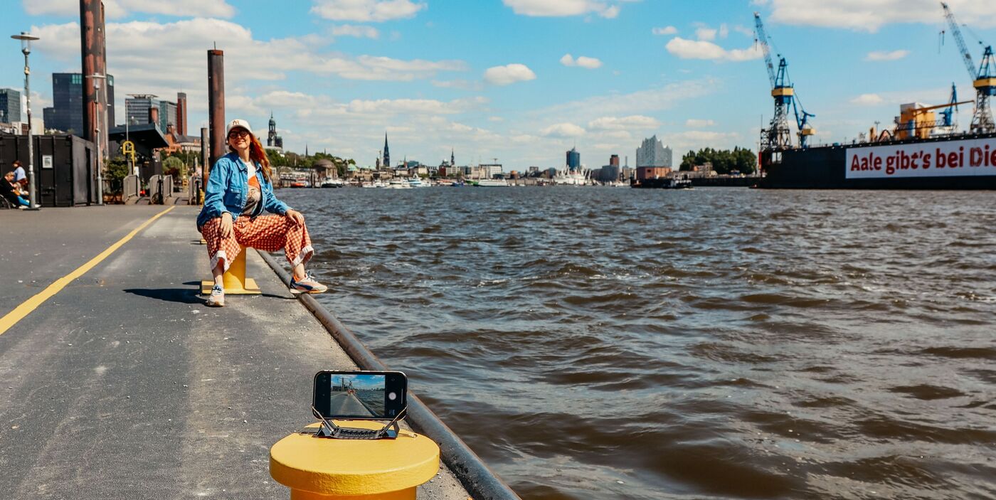 Person am Wasser sitzt auf einem Poller, sonniger Tag, Hamburg im Hintergrund, Handy mit Stativ vorne