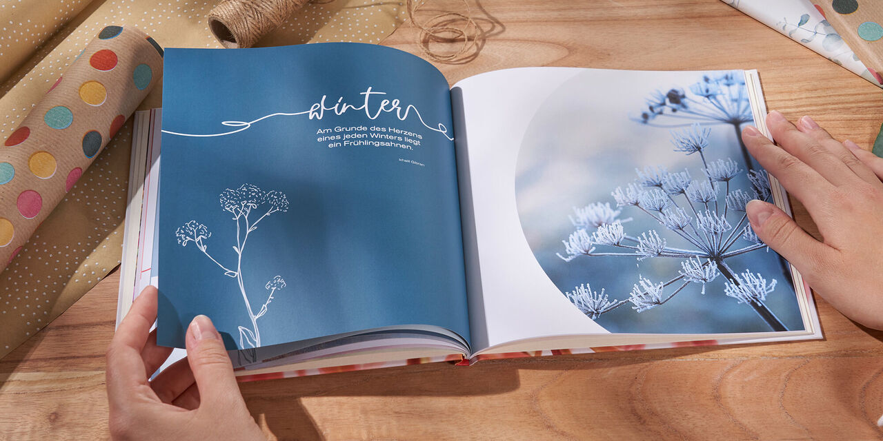 Zwei Hände blättern durch ein CEWE Fotobuch mit floralem Design und blauen Seiten.