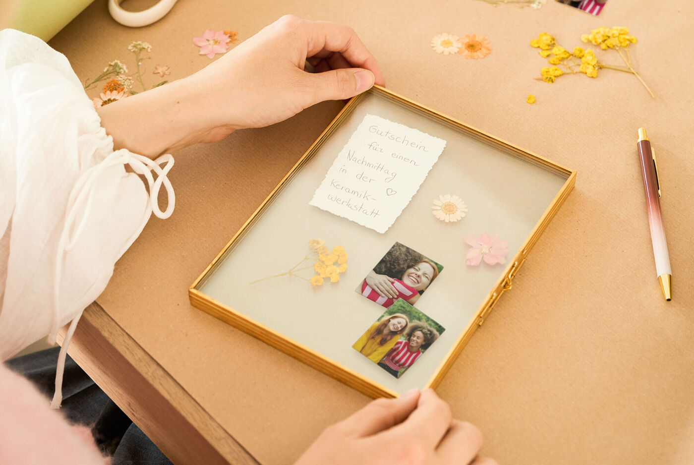 Hand legt einen handgeschriebenen Gutschein in einen goldenen Bilderrahmen, daneben getrocknete Blumen und zwei kleine Foto