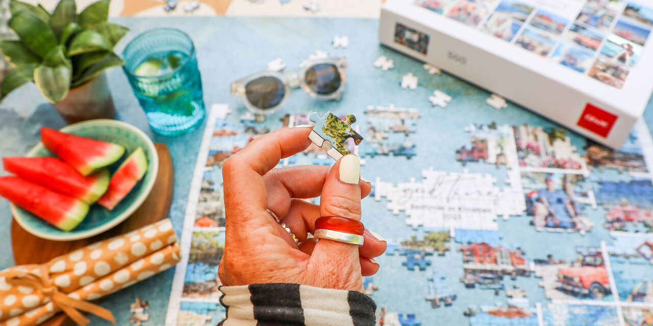 Eine Hand hält ein Puzzle-Stück mit einer Pflanze. Auf dem Tisch sind Wassermelonenscheiben, eine grüne Pflanze, Sonnenbrille, und ein CEWE Fotopuzzle zu sehen.