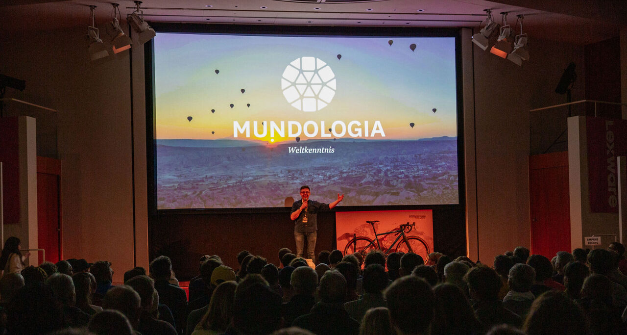 Ein Mann steht auf einer Bühne. Hinter ihm ist eine Leinwand, auf dem das Mundologia-Logo zu sehen ist.