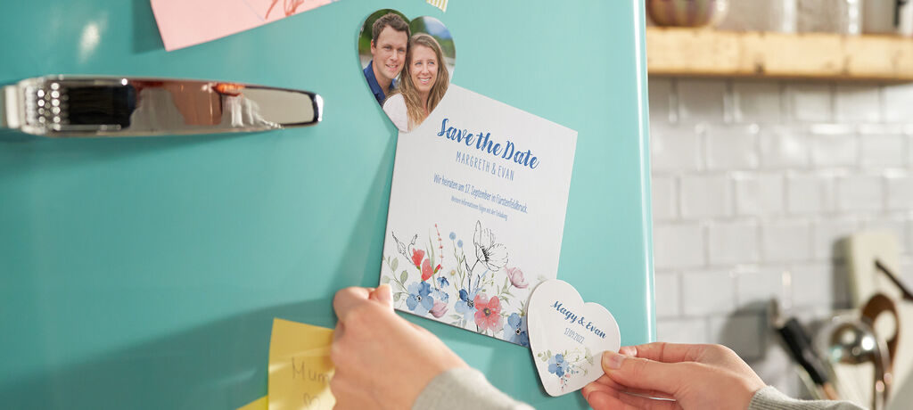 Eine Person klebt eine Einladung mit 'Save the Date' an den Kühlschrank. Links sind bunte Post-its, rechts sieht man zwei Herzen.