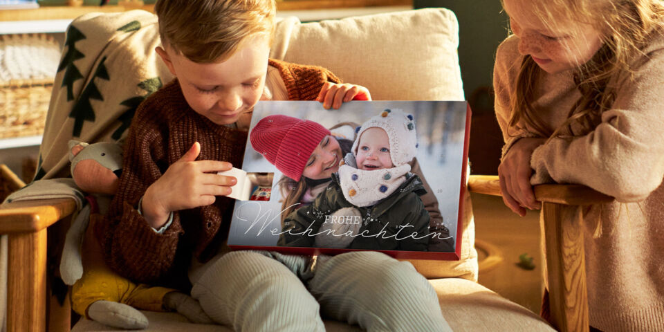 Zwei Kinder öffnen ein Türchen eines CEWE Adventskalenders mit Winterfoto und der Aufschrift „Frohe Weihnachten“ im warmen Licht