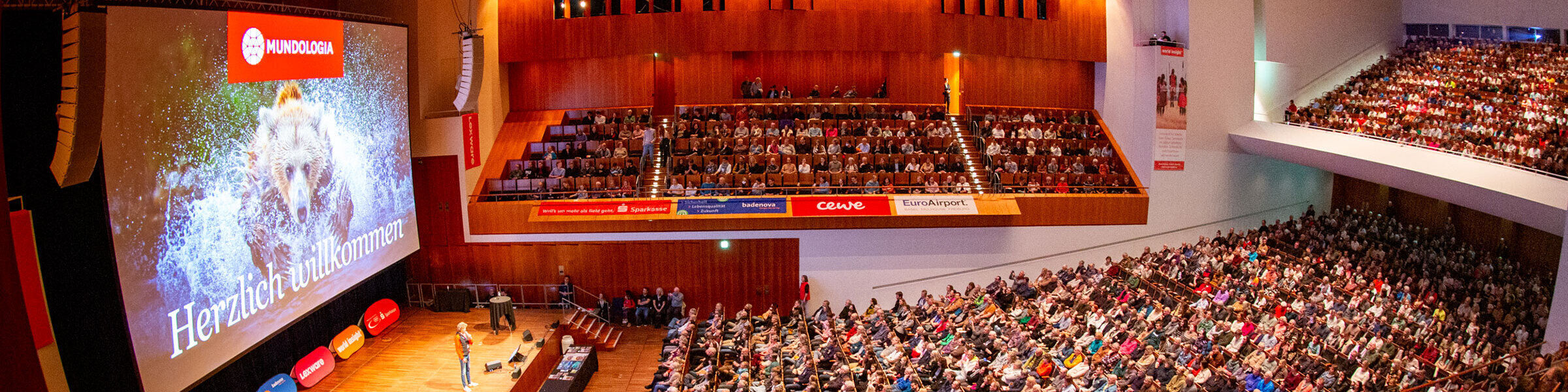 In einem Konzertsaal sitzen viele Menschen. Auf der Bühne steht ein Mann. Hinter ihm auf der Leinwand ist das Mundologia-Logo zu sehen.