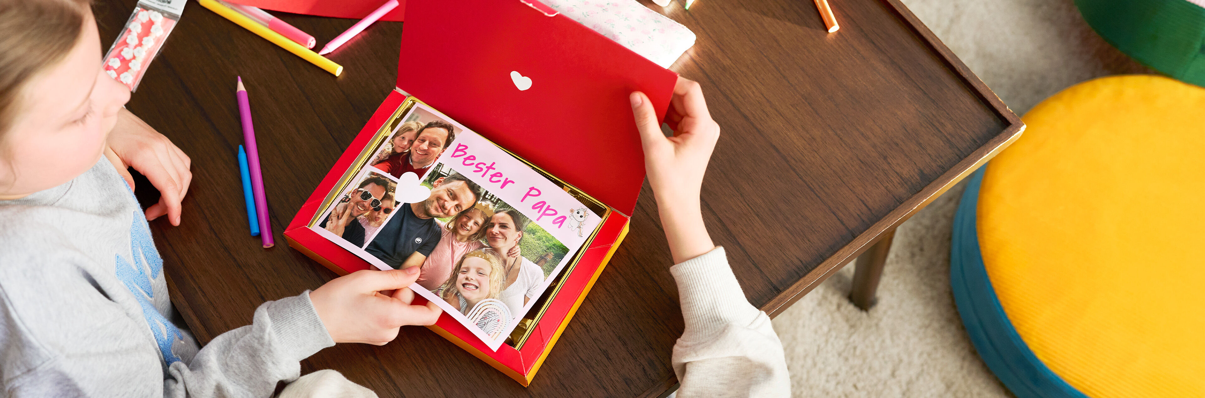 Ein Kind hält eine rote Pralinenschachtel mit einer personalisierten Collage auf der "Bester Papa" steht und Fotos sind.