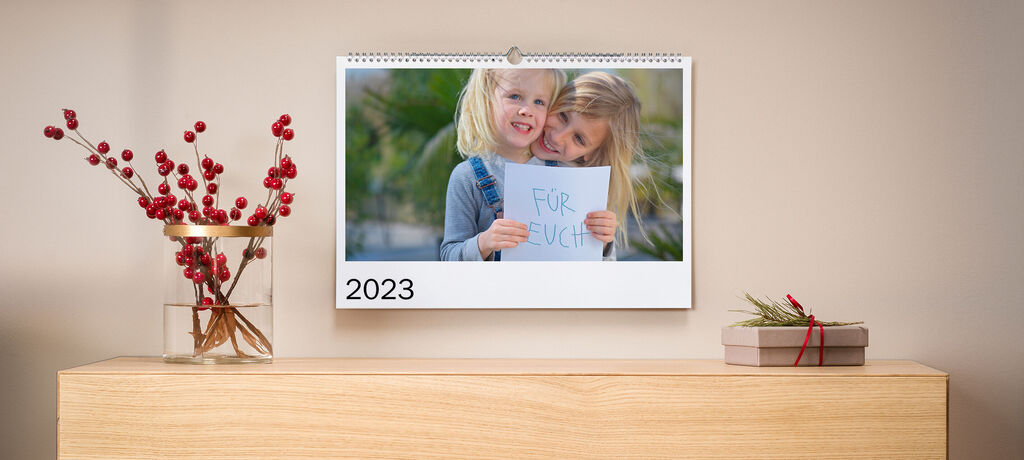 Zwei Personen halten ein Schild mit der Aufschrift 'Für euch' vor einem Kalender 2023, dekoriert mit einer Vase und Kräutern