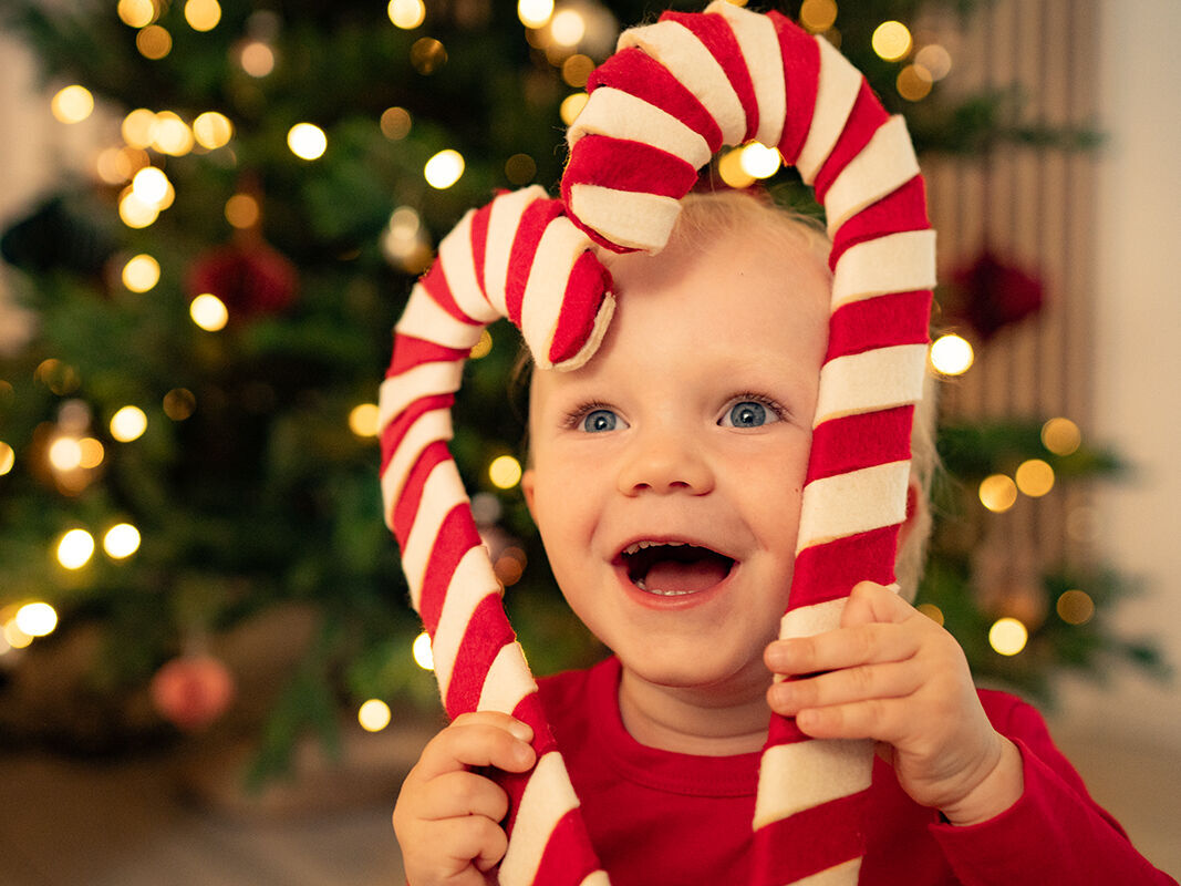 Ein Kind hält einen großen, rot-weißen Zuckerstangen-Herz vor einem festlich geschmückten Weihnachtsbaum.