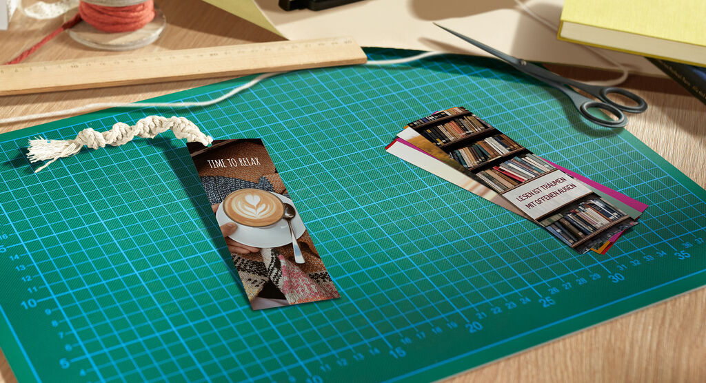 Zwei liebevoll gestaltete Lesezeichen liegen auf einem grünen Schneidebrett. Eines zeigt einen Kaffee, das andere hat ein Bücherregal abgebildet, ideal für Lesefans.