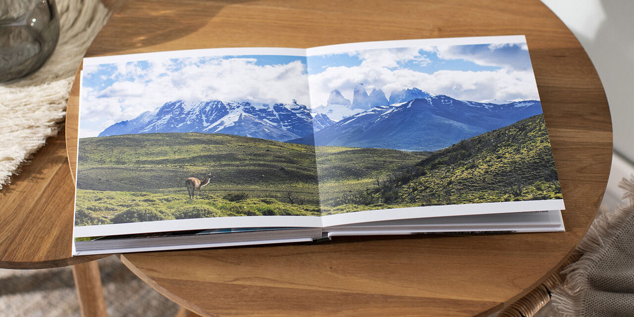 Ein offenes CEWE Fotobuch mit Naturbildern auf einem Tisch, im Hintergrund sind Berge und ein klarer Himmel zu sehen