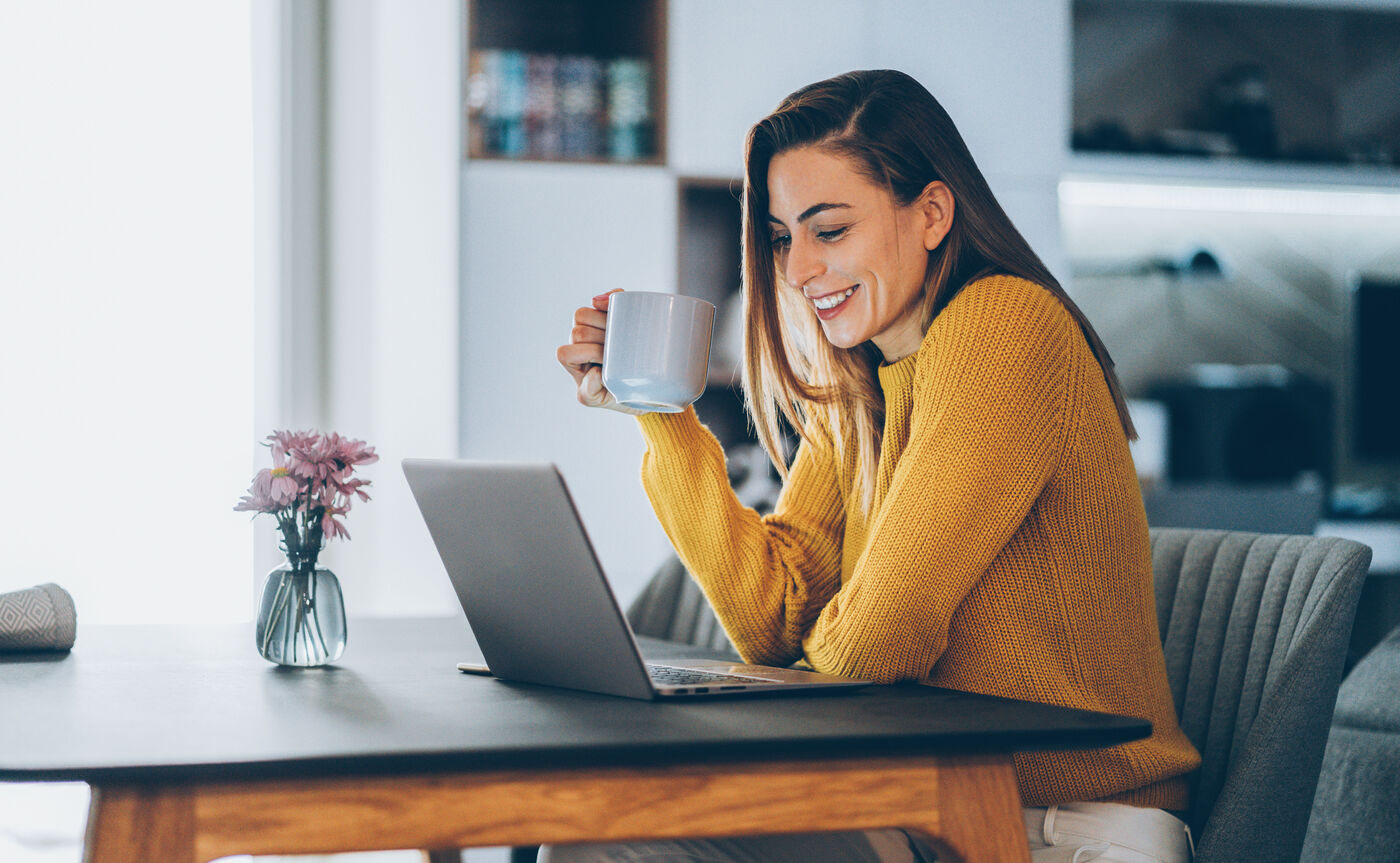Eine Frau sitzt an einem Tisch vor einem Laptop und lächelt. In der rechten Hand hält sie eine Tasse.