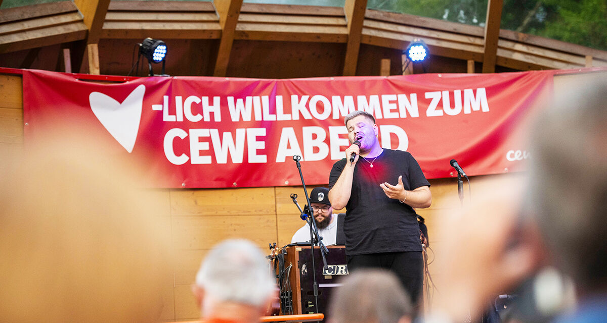 Ein Mann mit einem Mikrofon in der Hand steht vor einem Banner mit der Aufschrift "Herzlich willkommen zum CEWE Abend".