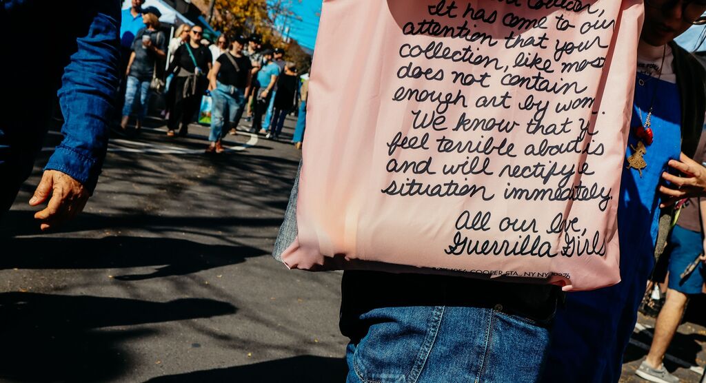 Person in Jeans trägt eine rosa Stofftasche mit schwarzer Schrift, im Hintergrund viele Menschen auf einer belebten Straße