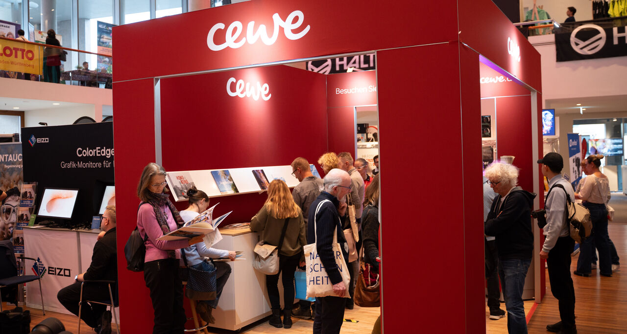 In einer Halle ist ein Messestand von CEWE aufgebaut. Viele Leute betrachten den Stand und die ausgelegten Fotobücher.