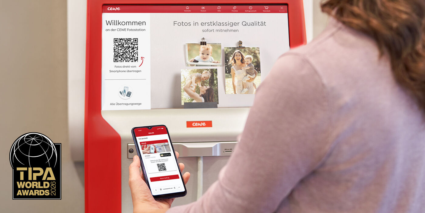 Person steht mit Handy in der Hand vor der CEWE Fotostation. Logo vom TIPA im Bild enthalten.