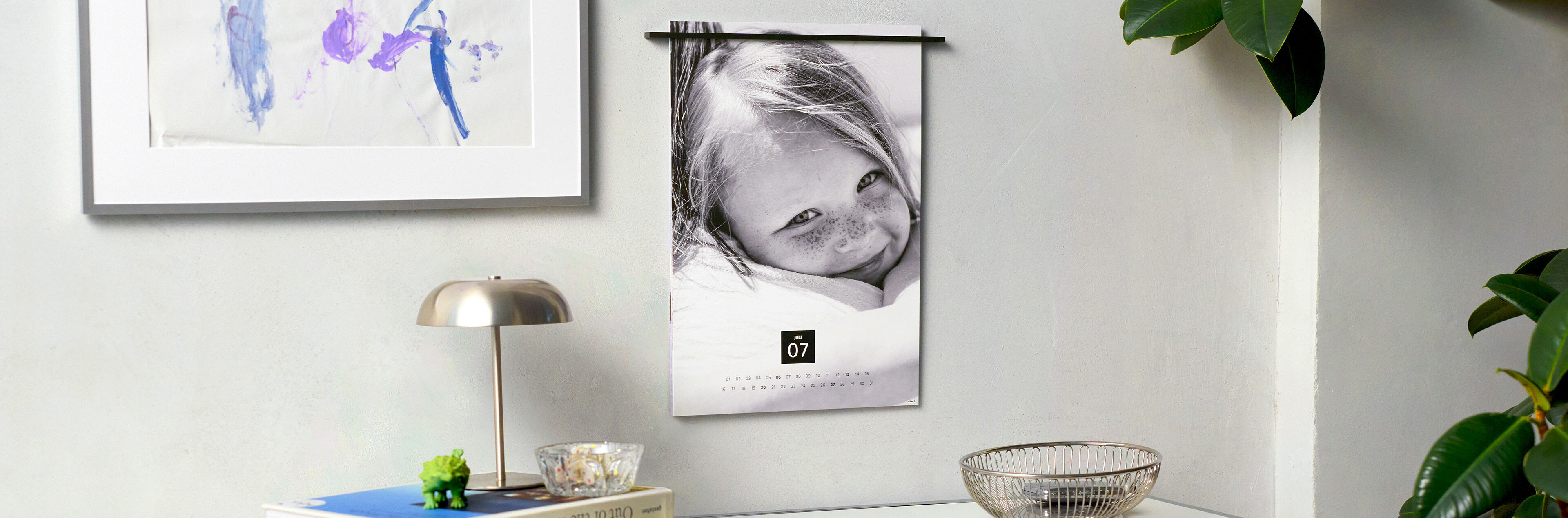 CEWE Fotokalender in schwarz-weiß hängt an einer hellen Wand über einem Schreibtisch mit Deko, Lampe und Pflanze.