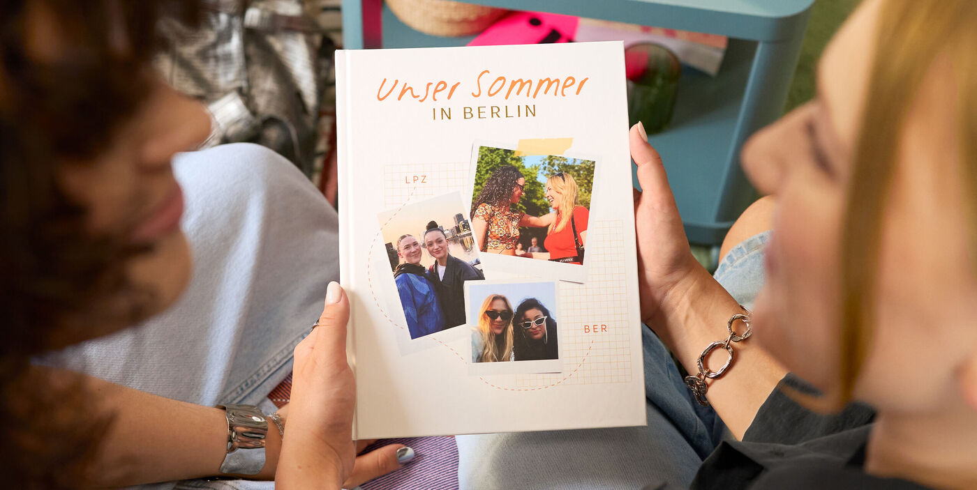 Zwei Frauen betrachten ein CEWE FOTOBUCH. Auf dem Cover sind drei Fotos von den beiden zu sehen.