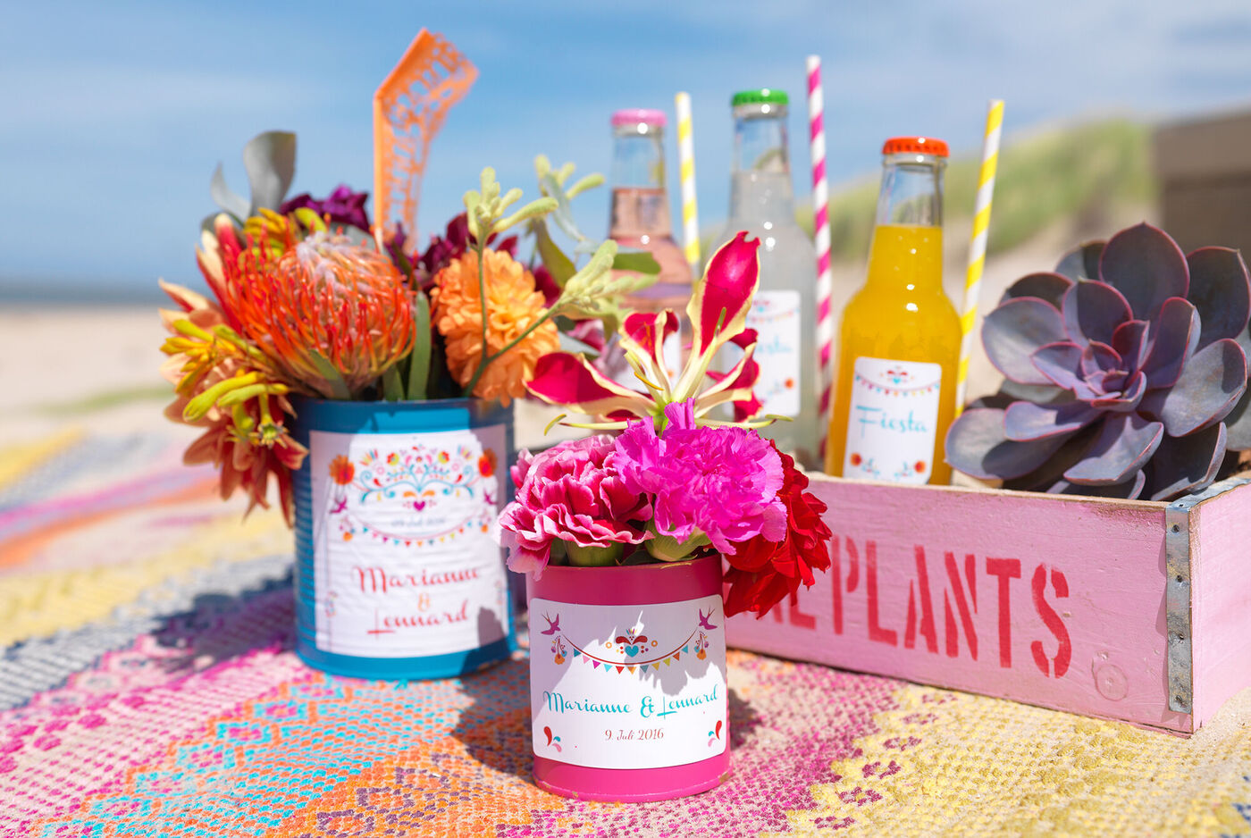 Bunte Blumenarrangements in Dosen auf einem bunten Teppich am Strand mit Erfrischungsgetränken