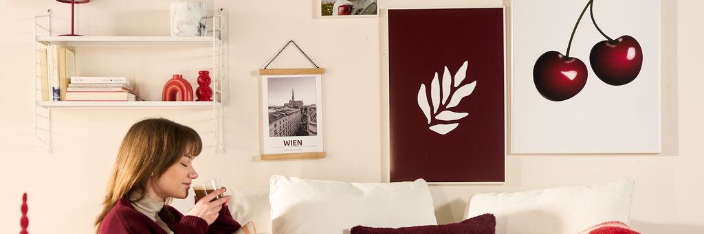 Eine Frau sitzt auf dem Sofa mit einem Getränk, rote Bilder und Poster hängen an der Wand.