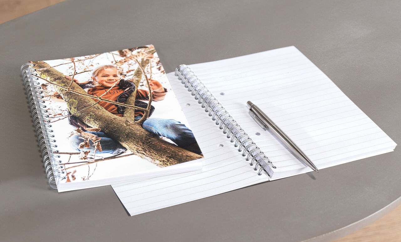 Ein CEWE Fotobuch auf einem Tisch mit einem Kinderfoto und ein leeres Notizbuch.