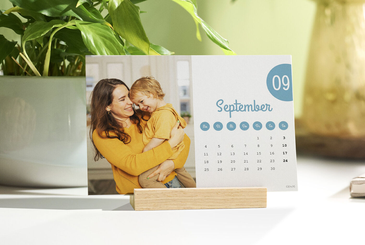 Ein CEWE Fotokalender im Holzständer zeigt auf der September‑Seite eine Mutter mit Kleinkind die sich liebevoll umarmen.