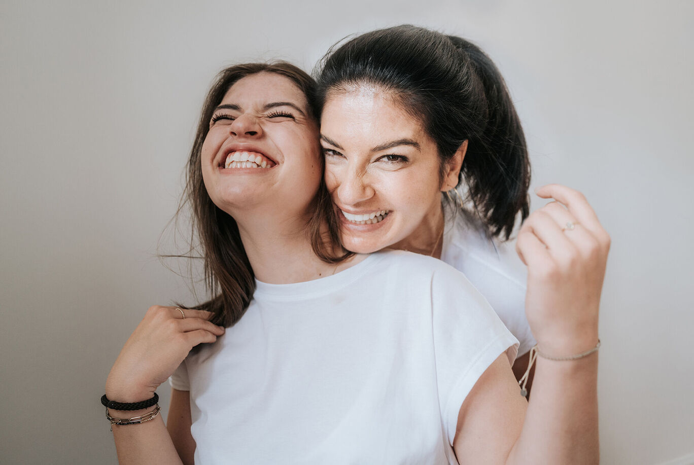 Zwei Frauen stehen nah beieinander und lächeln. Sie tragen weiße T-Shirts und zeigen Freude.