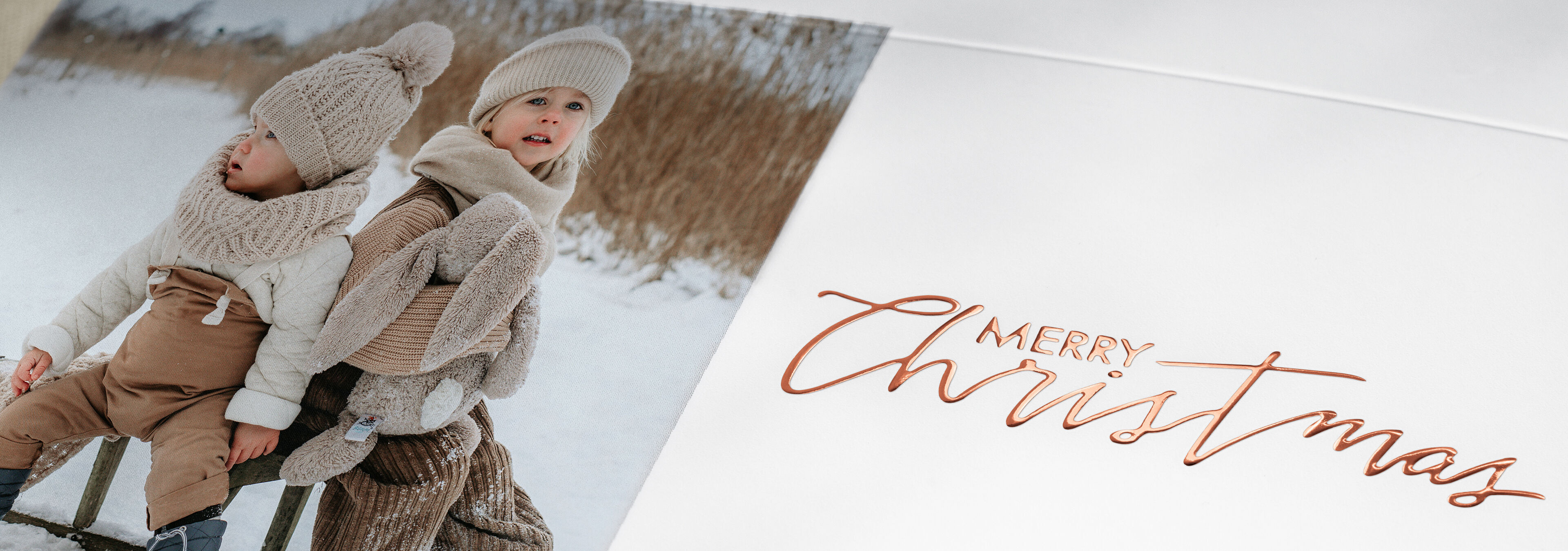 Zwei Kinder sitzen auf einer Bank in festlicher Winterkleidung, umgeben von Natur, mit einem 'Merry Christmas'-Text.