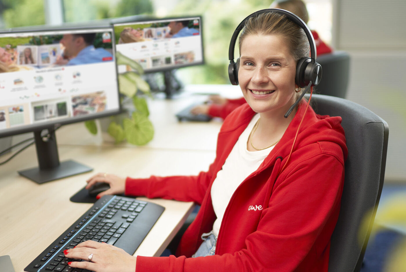 Kundenservice-Mitarbeiterin im roten CEWE Pullover mit Headset am Schreibtisch, arbeitet am Computer mit der CEWE Fotowelt