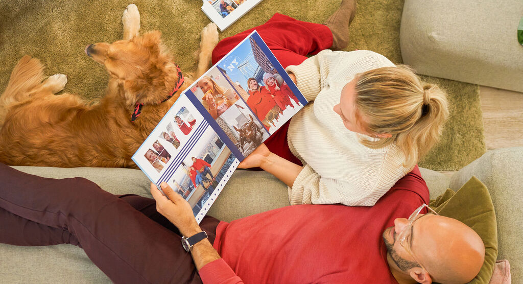 CEWE FOTOBUCH zeigt Reisebilder aus New York, zwei Menschen und ein Hund schauen begeistert hinein.
