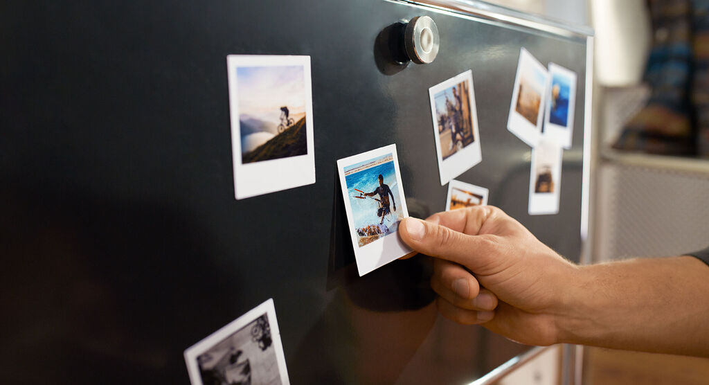 Eine Hand, die ein Polaroidfoto an einem Kühlschrank anbringt. Rundherum sind weitere Fotos befestigt.