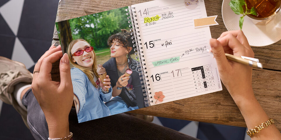 Ein geöffneter Terminplaner mit einem persönlichen Foto zweier Freundinnen liegt auf einem Holztisch, während eine Person darin schreibt.